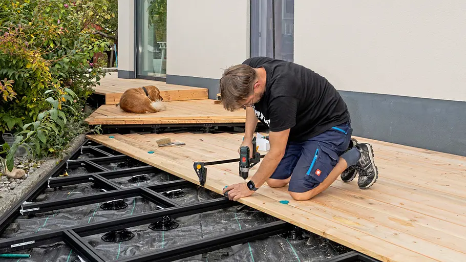 Terrasse gestalten Ein Mann befestigt Holzdielen auf einer Metallunterkonstruktion, während ein Hund auf dem fertigen Terrassenteil liegt.