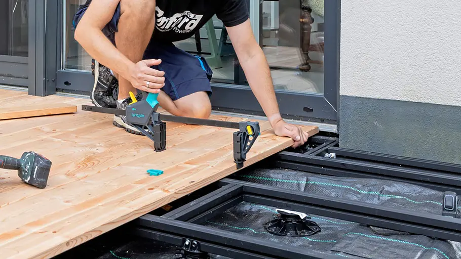 Terrasse gestalten Ein Mann zieht eine Holzdiele mit einer Zwinge gerade, bevor sie auf der Terrassenunterkonstruktion befestigt wird.