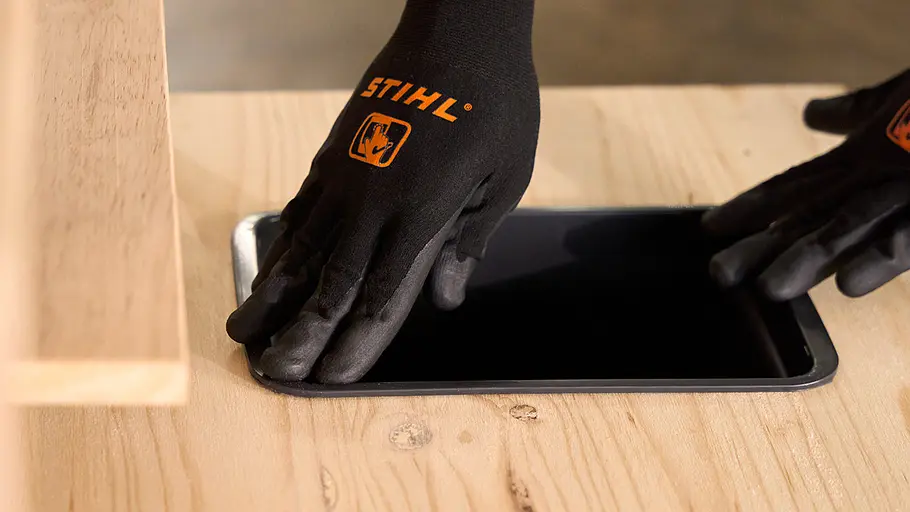Matschküche: Waschbecken einhängen Zwei Hände mit STIHL-Handschuhen setzen ein schwarzes Waschbecken in die ausgeschnittene Öffnung der Holzarbeitsplatte ein.