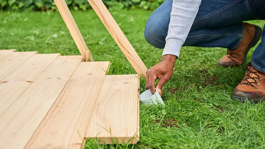 Eine Person kniet im Gras und platziert ein weißes Kunststoffrohr neben der Holzbodenplatte des Zeltes.