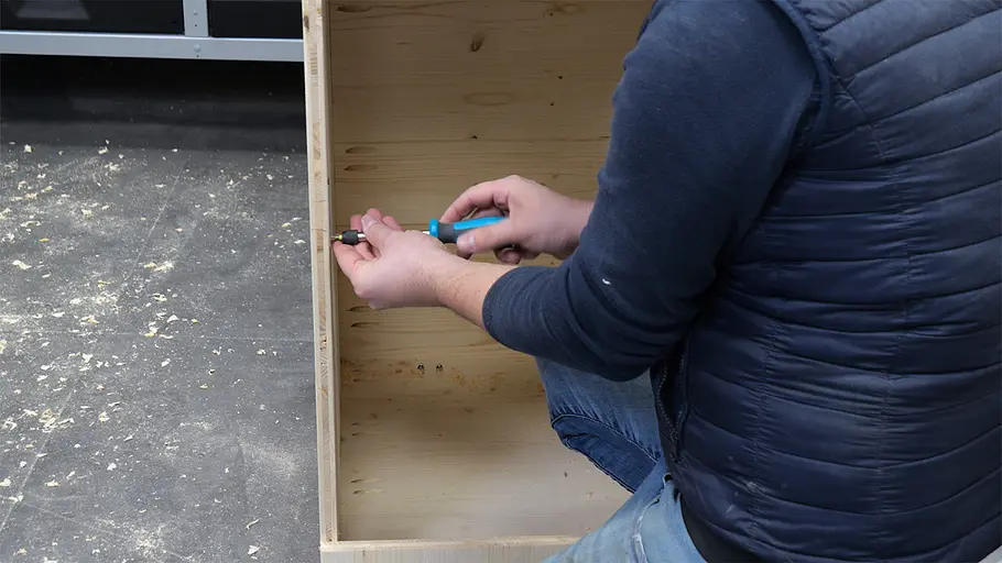 Paketbox Eine Person im blauen Pullover verschraubt mit einem Schraubendreher die Seitenwand einer hölzernen Paketbox auf dem Boden.