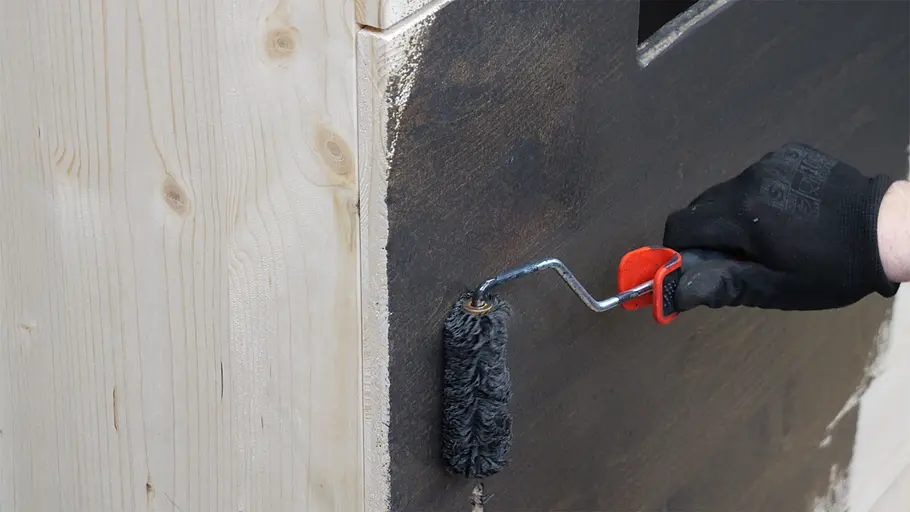 Paketbox Eine Person mit schwarzem Handschuh streicht die Außenseite einer hölzernen Paketbox mit einer kleinen Farbrolle dunkel an.