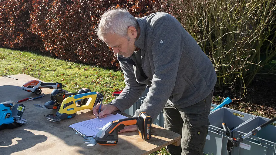 Akku-Grasscheren-Test: Dokumentation Ein Mann beugt sich über einen Tisch im Garten, schreibt Notizen und dokumentiert Testergebnisse mehrerer Akku-Grasscheren.