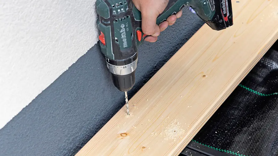 Terrasse gestalten Eine Hand bohrt mit einer Bohrmaschine Löcher in ein Holzbrett, das auf einer Terrassenunterkonstruktion liegt.