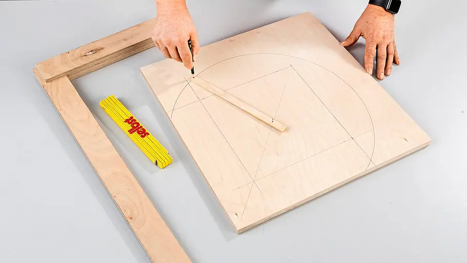 Linien anzeichnen Auf einer Holzplatte sind Linien und ein Viertelkreis aufgezeichnet, eine Hand benutzt einen Zirkel aus einer Leiste und einem Bleistift.