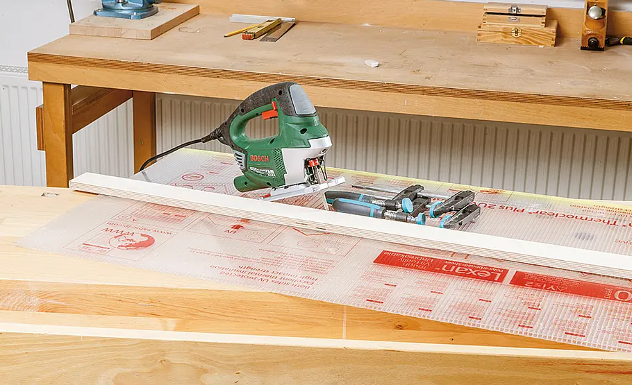 Eine Stichsäge liegt auf einer transparenten Kunststoffplatte, daneben Schraubzwingen und eine Holzleiste auf der Werkbank.