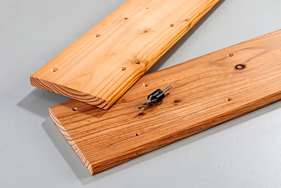 Gartendusche bauen Zwei zugeschnittene Holzbretter mit kleinen schwarzen Metallverbindern und Schrauben auf einer grauen Arbeitsfläche.