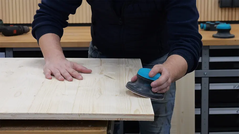 Paketbox Eine Person schleift die Oberfläche eines hellen Holzbretts mit einem elektrischen Exzenterschleifer ab.