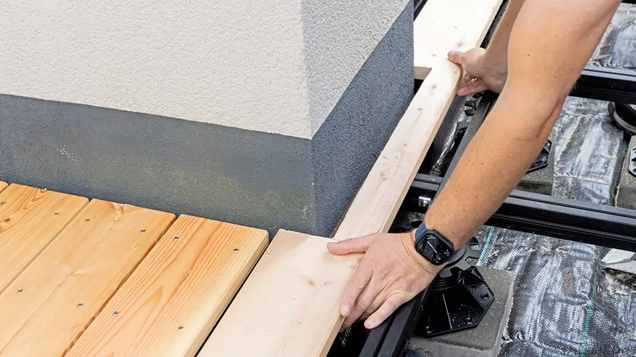 Terrasse gestalten Eine Person legt ein zugeschnittenes Terrassendielenbrett passgenau um eine Hausecke auf die Unterkonstruktion.