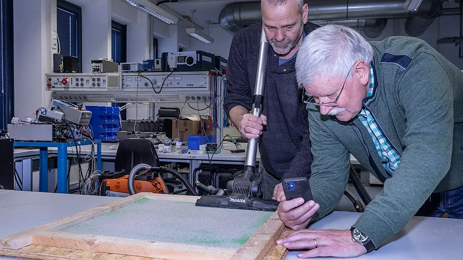 Teppich saugen Ein Mann saugt eine Platte mit einem Werkstattsauger ab, ein zweiter Mann hält ein Smartphone und beobachtet den Test.