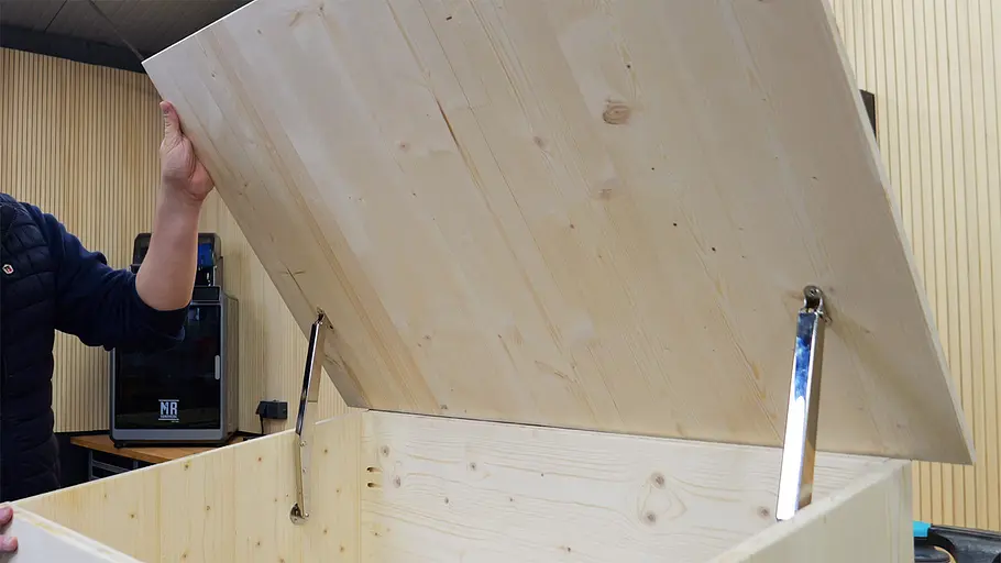 Paketbox Eine Person hebt den Deckel einer großen Holzkiste an, die mit zwei Metallaufstellern befestigt ist.