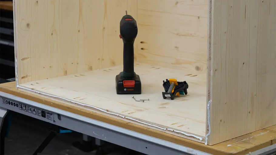 Paketbox Ein Akkuschrauber, einige Schrauben und eine kleine Zwinge liegen in einem teilweise zusammengebauten Holzkorpus auf einer Werkbank.