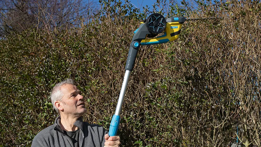 Akku-Grasscheren-Test: Kopfschnitt Ein Mann hält eine gelb-blaue Akku-Schere mit Teleskopstange, um eine hohe Hecke oberhalb seines Kopfes zu schneiden.