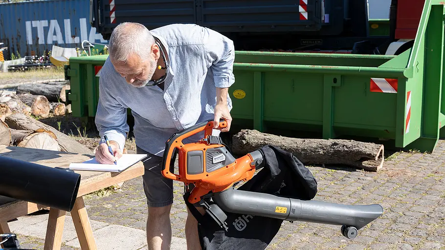 Ein Mann beugt sich über einen Holztisch, hält einen Laubsauger in der Hand und macht Notizen auf einem Block.