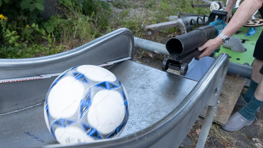Ein Laubsauger pustet einen Fußball auf einer Metallrutsche, die Szene wird aus der Nähe gezeigt.