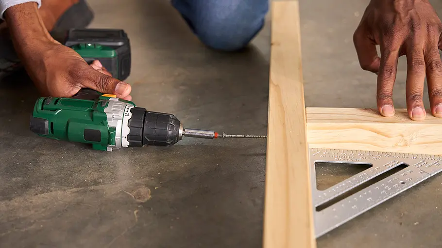 Matschküche: Verschrauben Eine Hand führt eine grüne Akkubohrmaschine, um zwei Kanthölzer mit einer Schraube auf einem Winkel zusammenzufügen.