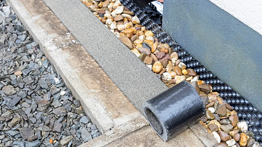 Terrasse gestalten Baustellendetail mit Kies, Splitt, Randsteinen, einer Gummibahnrolle und einer Noppenbahn an der Hauswand.