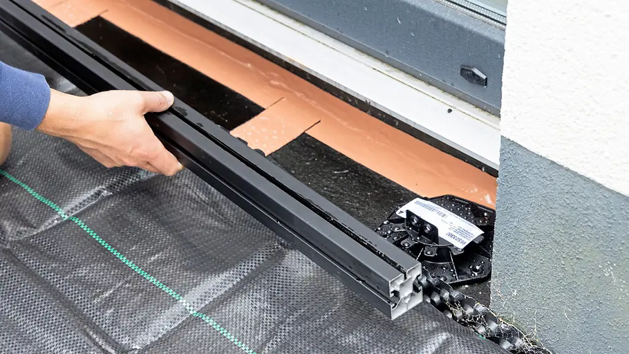 Terrasse gestalten Eine Hand platziert eine schwarze Metallkonstruktion auf einem Unkrautvlies direkt an der Terrassenkante vor dem Haus.