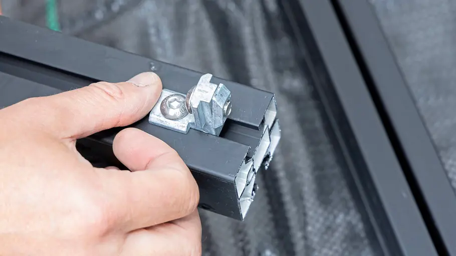 Terrasse gestalten Eine Hand hält ein schwarzes Aluminiumprofil, an dem ein Metallverbinder mit Schraube montiert ist, auf schwarzem Untergrund.