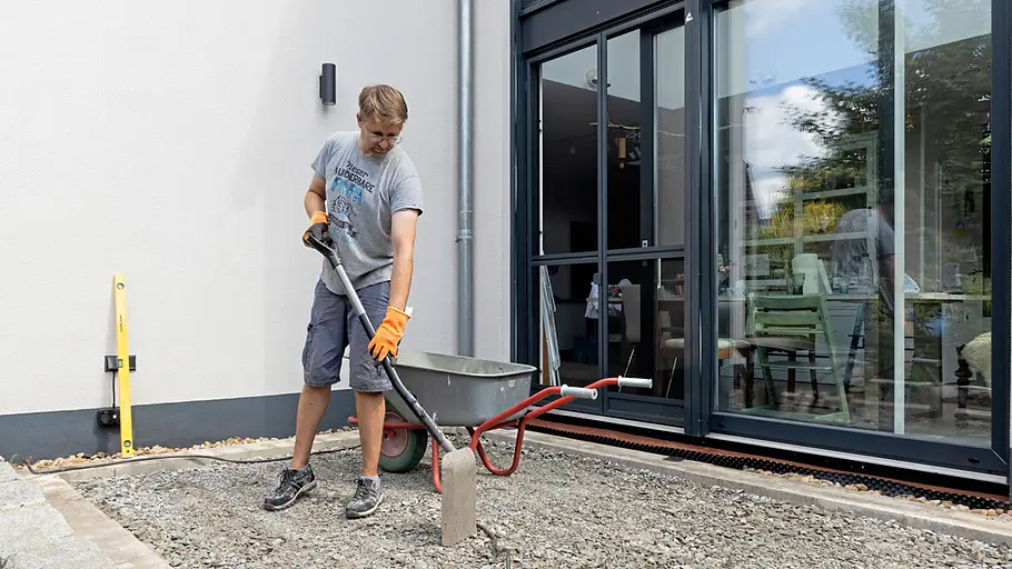 Terrasse gestalten Ein Mann bereitet im Freien mit Schaufel und Schubkarre den Untergrund vor einer Terrassentür für den Terrassenbau vor.