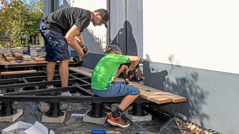 Terrasse gestalten Ein Erwachsener und ein Kind verschrauben gemeinsam Terrassendielen auf der Unterkonstruktion nahe der Hauswand.
