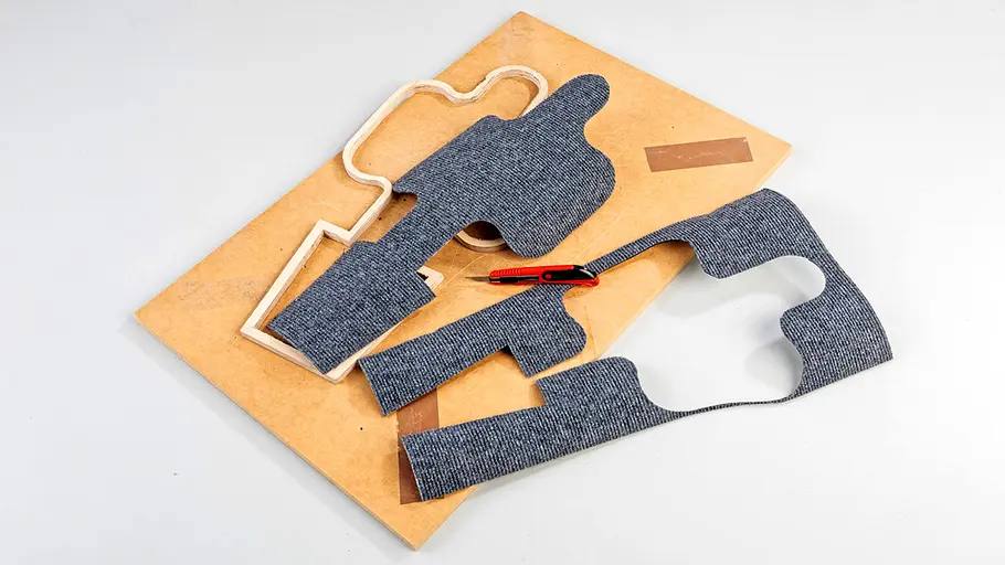 Kratzmatte zuschneiden Ein Stück grauer Teppich wurde in Kaktusform ausgeschnitten, die Einzelteile und ein Cutter liegen auf einer Holzplatte.