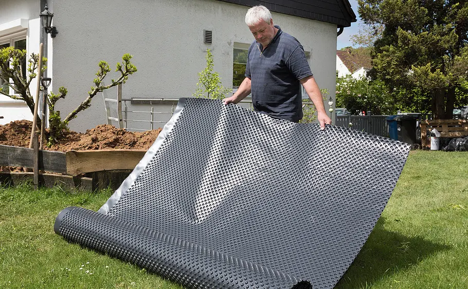 Ausrollen einer Noppenfolie zur Außenwandabdichtung Ein Mann steht auf einer Wiese und rollt eine große Noppenfolie vor einem Haus aus.