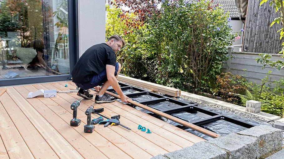 Terrasse gestalten Ein Mann legt weitere Holzdielen für die Terrasse bereit, das bereits verlegte Holz ist teilweise fertiggestellt.
