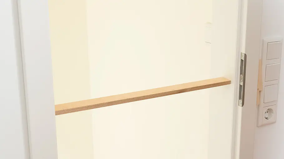 Türzargen spreizen Eine Tür mit weißem Türrahmen, innen quer eine Holzleiste eingespannt, daneben Lichtschalter und Steckdose.