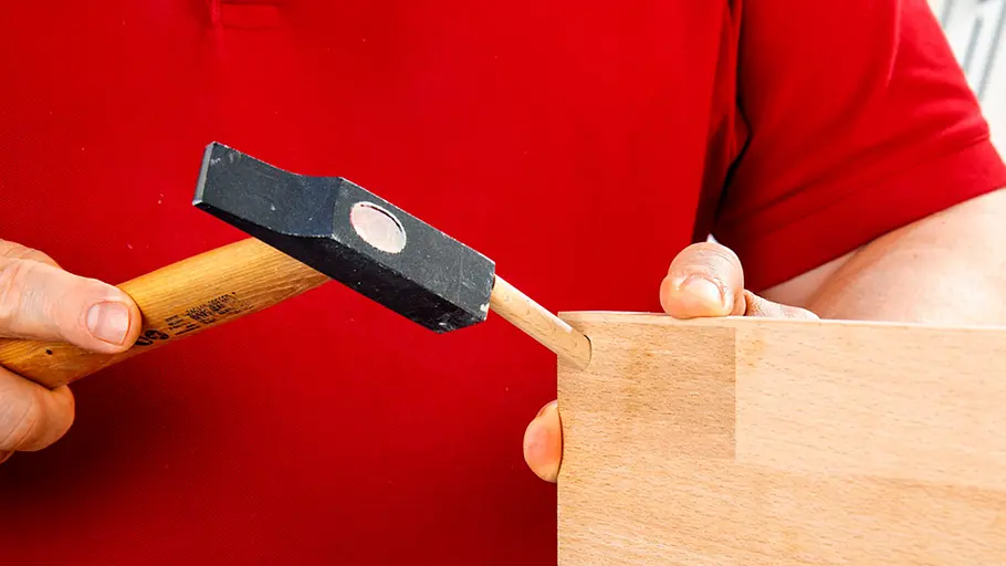 Verdrehter Hocker Ein Holzrundstab wird mit einem Hammer in eine gebohrte Öffnung an der Kante eines Holzbretts eingetrieben.
