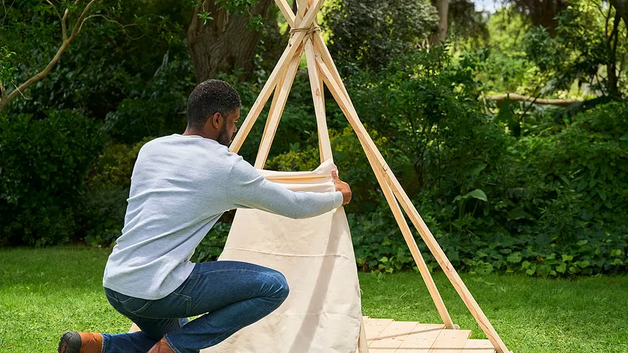 Ein Mann kniet vor dem Tipi-Gestell und befestigt einen großen Stoff an den Holzlatten des Zeltes im Garten.