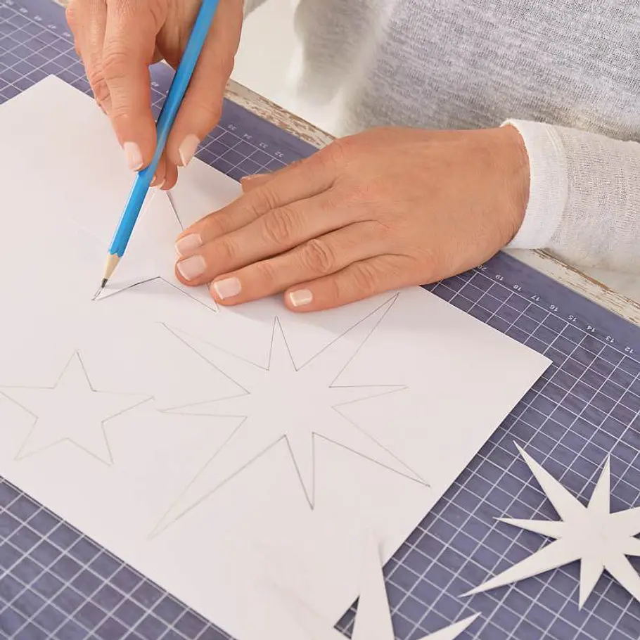 Weihnachts-Windlicht: Funkelndes Sternen-Glas Die Hand einer Person zeichnet Sterne auf weißes Papier für eine weihnachtliche Bastelarbeit.