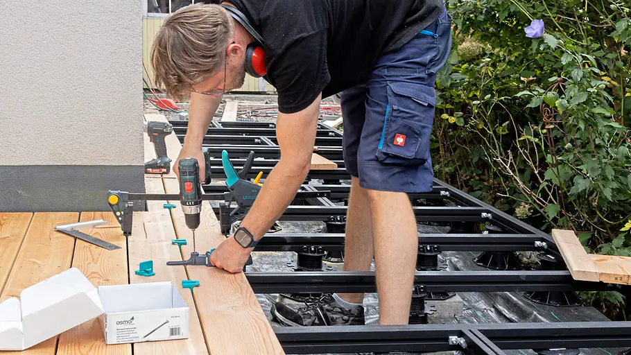Terrasse gestalten Ein Mann verschraubt mit einem Akkuschrauber Terrassendielen, die mit Abstandshaltern auf der Unterkonstruktion fixiert sind.