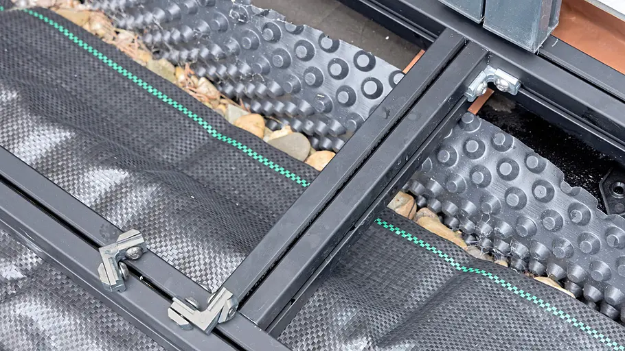 Terrasse gestalten Schwarze Metallunterkonstruktion mit Montageschienen und Verbindern liegt auf Unkrautvlies, Kies und Noppenfolie an der Hauswand.