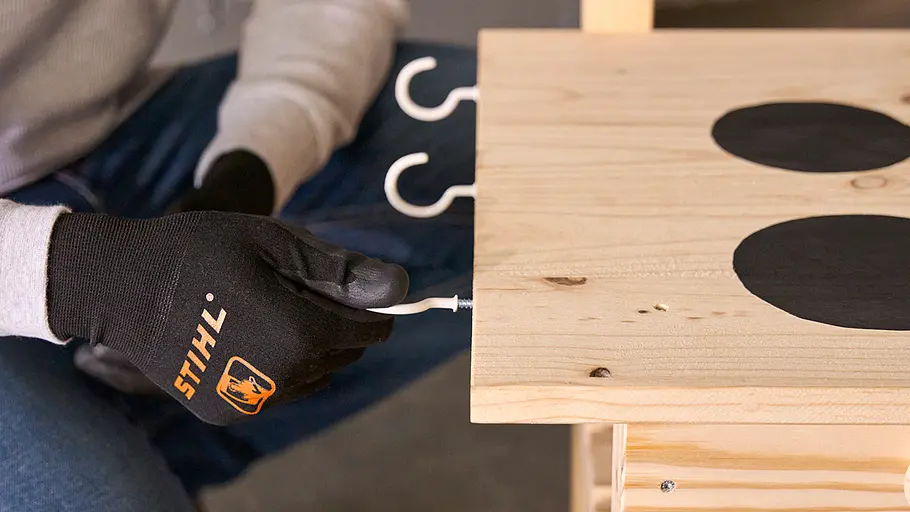 Matschküche: Schraubhaken Eine Person mit STIHL-Handschuhen schraubt mehrere weiße Schraubhaken an die Seitenkante der Arbeitsplatte aus Holz.