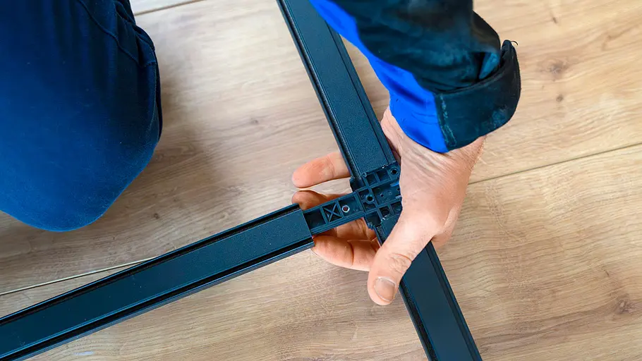 Eine Person setzt auf einem Holzfußboden einen schwarzen Metallrahmen mit Eckverbinder für eine Insektenschutztür zusammen.