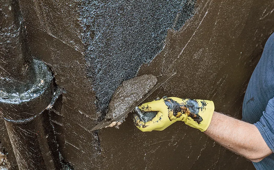 Hauswand mit Bitumen abdichten Eine Person trägt mit gelbem Handschuh Bitumen mit einer Kelle auf eine Hauswand auf.