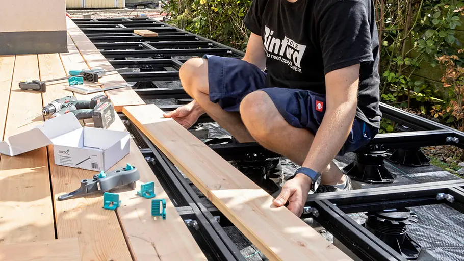 Terrasse gestalten Ein Mann legt eine Holzdiele auf die Metallunterkonstruktion der Terrasse und richtet sie für die Montage aus.