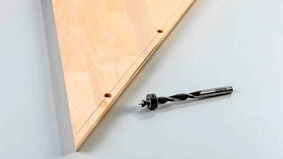 Verdrehter Hocker Ein Holzbrett mit zwei gebohrten Löchern liegt auf einer Fläche, daneben liegt ein Metallholzbohrer.