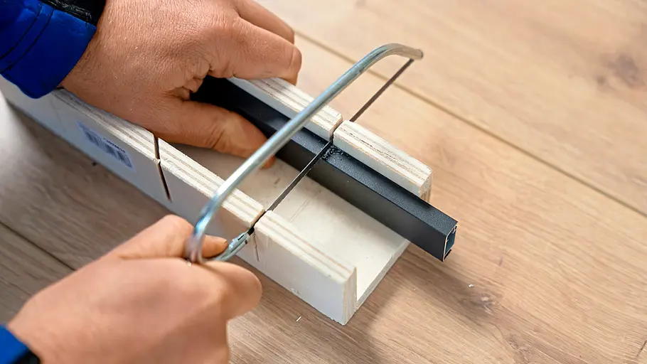 Mit einer Metallsäge wird ein schwarzes Aluprofil auf einer Schneidlade auf einem Holzboden zugeschnitten.