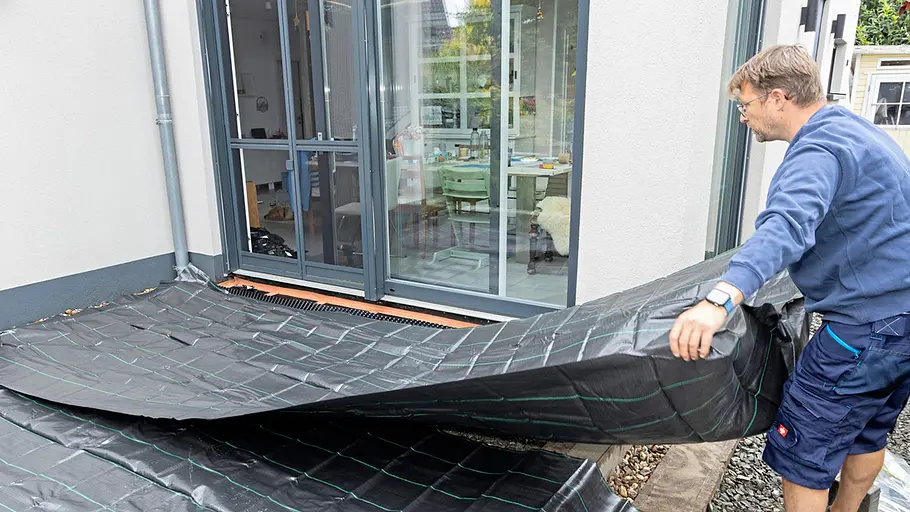 Terrasse gestalten Ein Mann breitet vor der Terrassentür ein großes schwarzes Unkrautvlies auf dem vorbereiteten Untergrund aus.