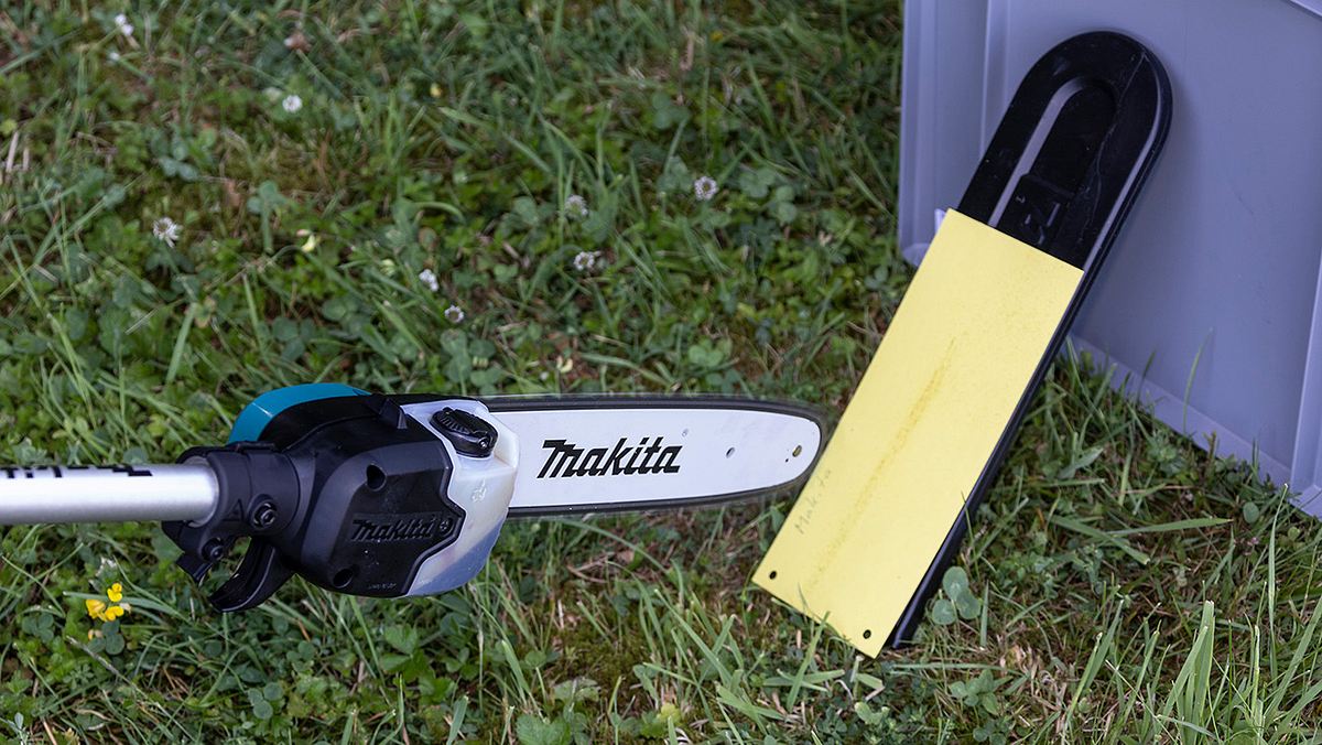 Eine Akku-Kettensäge von Makita liegt auf einer Wiese neben einem gelb-schwarzen Schutzkappe und einer grauen Kiste.