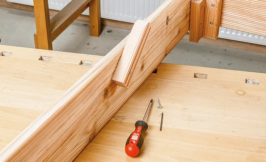 Ein Schraubendreher und Schrauben liegen auf einer Werkbank, ein Holzrahmen mit angeschraubtem Holzstück ist zu sehen.