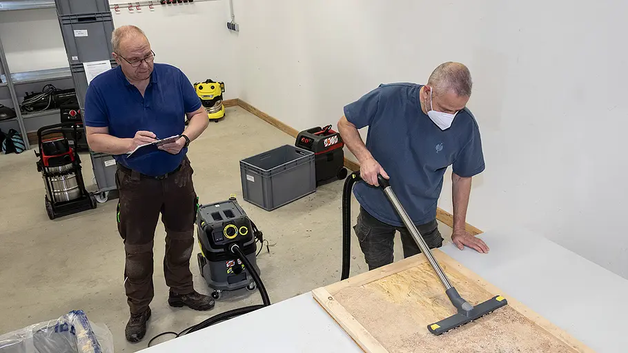 Randnah saugen Ein Mann mit Maske saugt Holzspäne von einer Arbeitsplatte, ein weiterer Mann macht Notizen in einer Werkstatt.