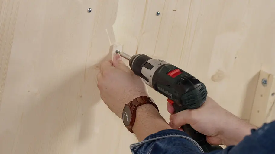 Paketbox Eine Person verschraubt mit einem Akkuschrauber eine Holzleiste an die helle Innenwand der Paketbox.