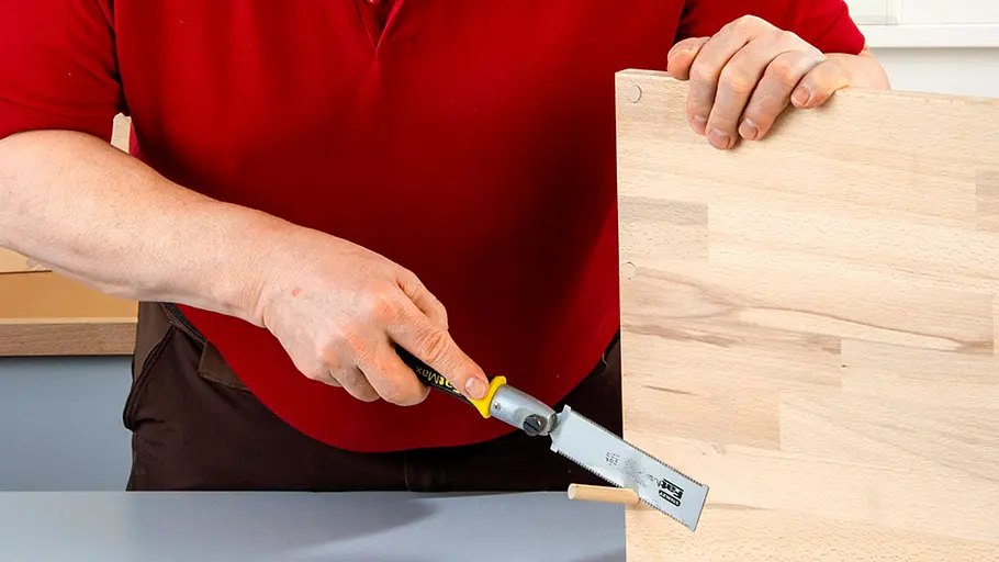 Verdrehter Hocker Ein Mann sägt mit einer Feinsäge einen überstehenden Holzdübel bündig an einer hellen Holzplatte ab.