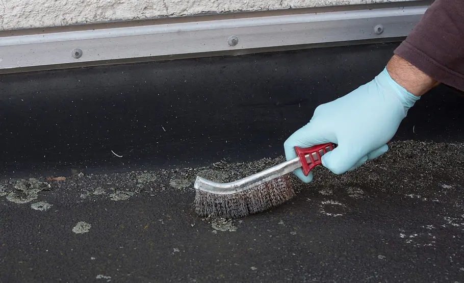 Mit einer Drahtbürste entfernt ein Handwerker Schmutz und Schadhaftes von einem Bitumendach