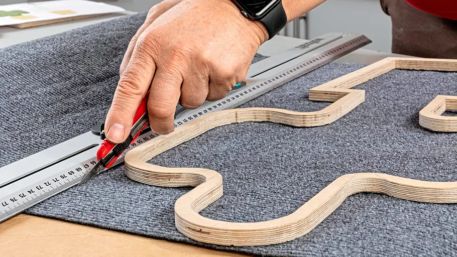 Kratzmatte zuschneiden Eine Hand schneidet mit einem Cutter und Lineal grauen Teppich entlang einer Sperrholz-Kaktusform für das Kratzbrett.
