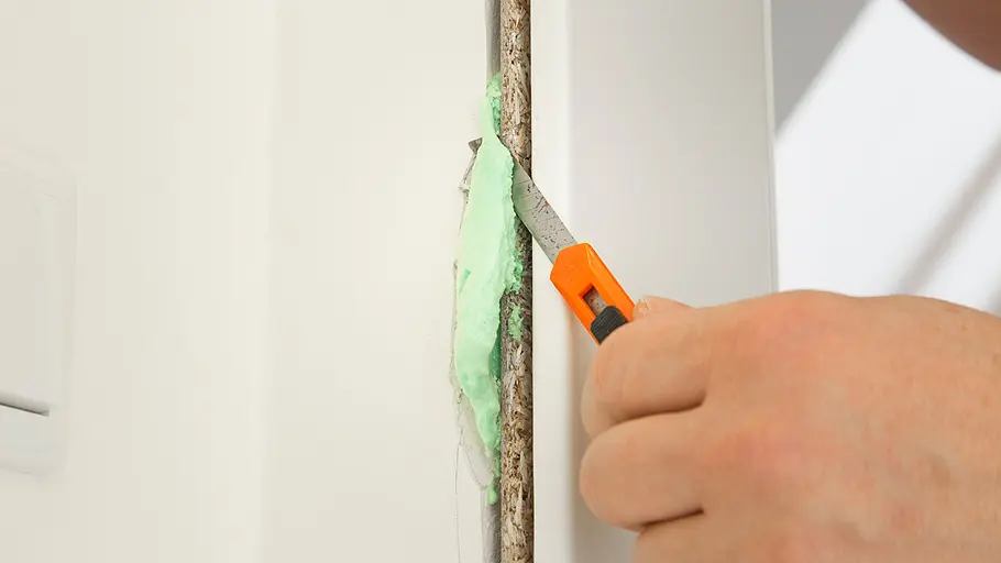 Schaumüberstände abschneiden Eine Hand schneidet mit einem Cuttermesser überschüssigen grünen Bauschaum am Türrahmen ab.