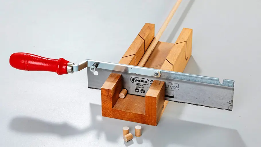 Verdrehter Hocker Eine kleine Handsäge mit Holzgriff liegt auf einer Gehrungslade, daneben mehrere abgesägte Holzdübelstücke.